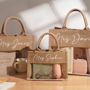 Peut inclure: Trois sacs fourre-tout rectangulaires en toile de jute avec des anses en tissu marron et des fenêtres en plastique transparent. Chaque sac porte une inscription blanche en cursive : Mrs. Jones, Mrs. Sophia ou Mrs. Davis, contenant divers objets.