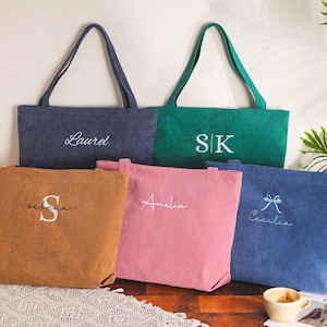 May include: Five personalized tote bags in various colors: navy, green, brown, pink, and blue. Each bag features embroidered text, including names and initials. The bags have long handles and a simple, rectangular design.