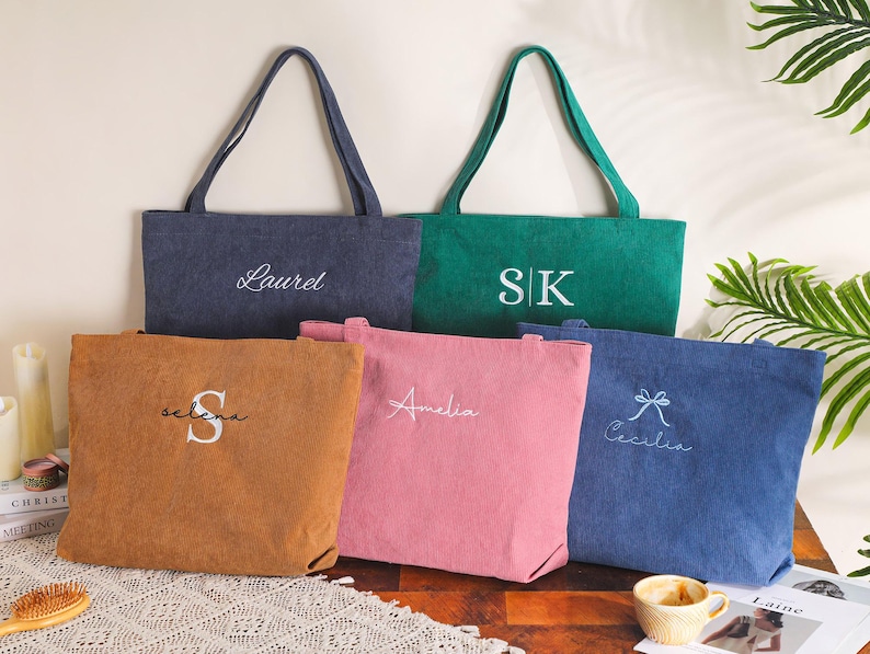 May include: Five personalized tote bags in various colors: navy, green, brown, pink, and blue. Each bag features embroidered text, including names and initials. The bags have long handles and a simple, rectangular design.