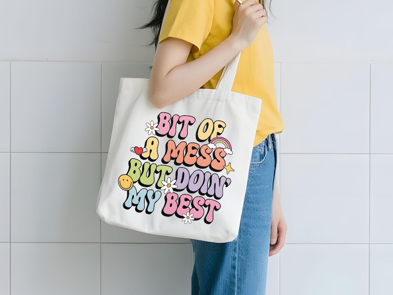 May include: Cream-colored tote bag with the text "bit of a mess but doin' my best" in colorful, retro-style lettering. The design includes a smiley face, rainbow, and flowers. The bag is held by a person wearing a yellow shirt and blue jeans.