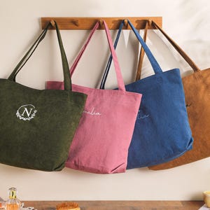 May include: Four corduroy tote bags in olive green, pink, blue, and tan hang on a wooden rack. Each bag has a long strap and a unique embroidered design. The olive green bag features a white "N" in a wreath.