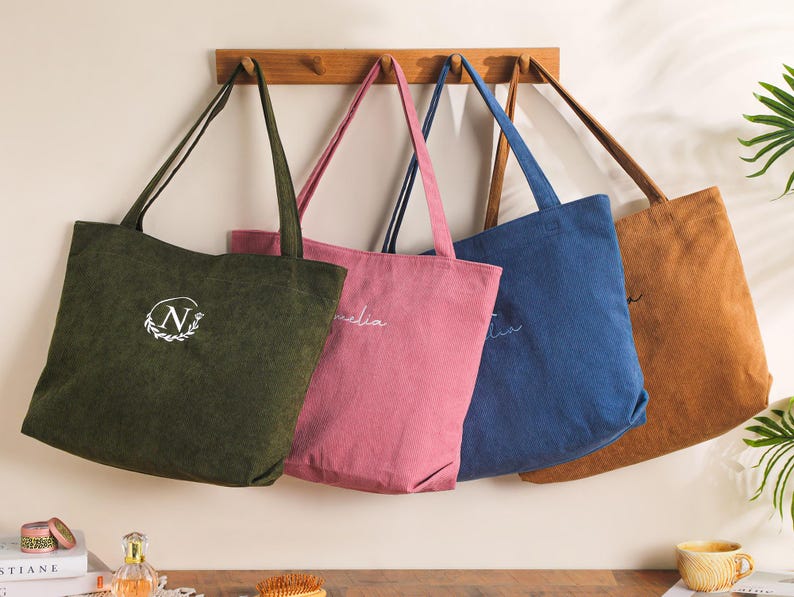 May include: Four corduroy tote bags in olive green, pink, blue, and tan hang on a wooden rack. Each bag has a long strap and a unique embroidered design. The olive green bag features a white "N" in a wreath.