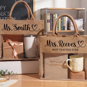 May include: Two rectangular jute tote bags with brown fabric handles and a clear plastic front window. The left bag features the text Ms. Smith with a heart icon. The right bag displays Mrs. Reeves and the text BEST TEACHER EVER with a heart icon.