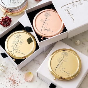 May include: Three round metal compact mirrors in gold and rose gold finishes, each featuring a unique floral line art engraving and a name: Belle, Sophia, Selena, and Emily. The mirrors are presented in individual white gift boxes.