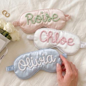 Puede incluir: Tres antifaces de satén para dormir con bandas elásticas. El antifaz superior es rosa claro con el nombre Roise en hilo verde. El antifaz central es blanco con el nombre Chloe en hilo rosa. El antifaz inferior es azul claro con el nombre Olivia en hilo blanco.
