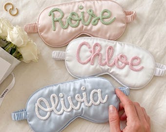 May include: Three satin sleep masks with elastic headbands. The top mask is light pink with the name Roise in green yarn. The middle mask is white with the name Chloe in pink yarn. The bottom mask is light blue with the name Olivia in white yarn.