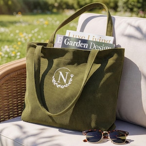 May include: Olive green corduroy tote bag with a white monogram "N" and laurel wreath design. The bag contains magazines with the text "Luxe Living" and "Garden Design". Tortoiseshell sunglasses rest on the surface. The bag is on a wicker chair.