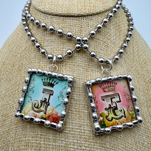 May include: Two silver-toned necklaces with square pendants. Each pendant features a letter, a crown, and floral accents. One pendant has a blue background, the other a pink background. The necklaces are displayed on a tan surface.