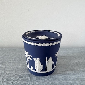 May include: A dark blue ceramic container with a lid. The container features white relief designs of figures and trees around the sides. The lid has a white relief design and a decorative border. The container is on a gray surface.
