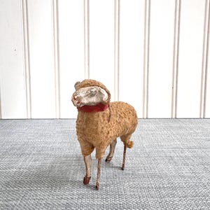 May include: A vintage toy sheep with a tan body, a white face, and a red collar. The sheep has thin, metal legs and a small, fluffy tail. The background is a light gray surface and a white wall.