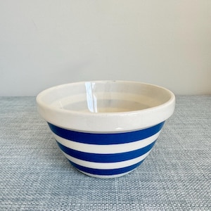 May include: A ceramic mixing bowl with a white rim and blue and white horizontal stripes. The bowl is round and has a classic design, suitable for kitchen use. The bowl is sitting on a blue surface.