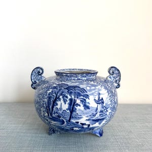 May include: A blue and white ceramic bowl featuring a transferware landscape pattern with trees, a windmill, and grazing cattle. The bowl has a rounded body, a wide circular opening, two ornate scroll-shaped handles, and three small feet.
