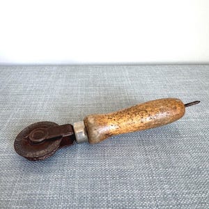May include: A vintage brown and tan seam roller tool with a wooden handle and a metal wheel. The tool has a metal pin extending from the end of the handle. The wheel is attached to a metal frame.