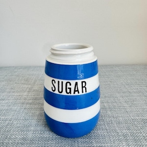 May include: A ceramic sugar jar with a blue and white striped pattern. The word "SUGAR" is printed in black on a white stripe. The jar has a white rim and a rounded base.