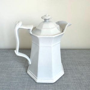 May include: A white ceramic coffee pot with a lid and handle. The pot has a geometric, octagonal shape and a decorative handle. The lid is topped with a small, floral-shaped knob. The pot is sitting on a gray surface.