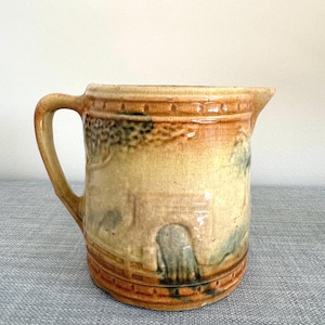 May include: A vintage ceramic pitcher with a handle and spout. The pitcher features a landscape design in shades of tan, brown, and green. The pitcher has a textured surface and a decorative rim. The pitcher is likely used for serving beverages.