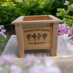 以下が含まれることがあります： 「Grammy Dot's Garden」の文字と花のイラストが描かれた、明るい色の木製プランターボックス。 「love grows here」というフレーズも刻まれています。 プランターは正方形で、4本の脚があり、木製の表面に置かれています。