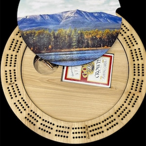 Op de afbeelding: Een houten cribbage-bord met een ronde, afneembare bovenkant met een schilderachtig berglandschap. Het bord is dubbelzijdig en bevat een kaartspel.