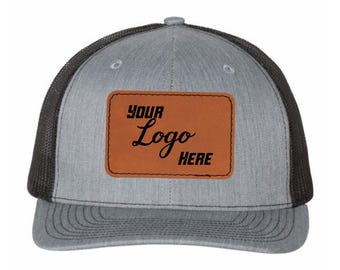 Könnte beinhalten: Graue Trucker-Kappe mit schwarzem Mesh-Rücken und gebogenem Schirm. Die Kappe hat einen braunen Lederaufnäher mit den Worten "YOUR Logo HERE" in schwarzer Schrift.
