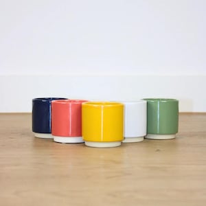May include: A set of five cylindrical ceramic cups arranged in a row, featuring a smooth, glossy finish in navy blue, coral, yellow, white, and sage green. Each cup has a distinct white base, creating a uniform, stackable design with clean lines.