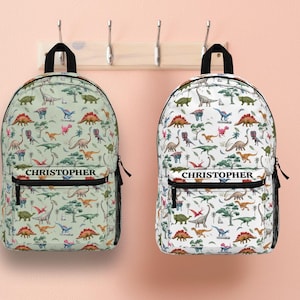May include: Two backpacks with a dinosaur pattern, one on a light green background and the other on a white background. Each backpack has the name "Christopher" printed on the front. The backpacks are hanging on a wooden rack.