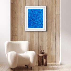 Puede incluir: Una pintura abstracta enmarcada en blanco con un fondo azul. La pintura está colgada en una pared de madera con un sillón blanco y una pequeña mesa auxiliar de madera delante.