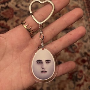 Può includere: Un portachiavi in acrilico trasparente con un anello e una catena a forma di cuore argentato. Il portachiavi presenta un ciondolo a forma di uovo con un'immagine stampata del volto di una persona. Il volto ha la pelle chiara e i capelli scuri.