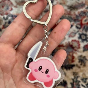 May include: A pink Kirby character keychain with a silver heart-shaped keyring. The character is holding a knife. The keychain is made of clear acrylic.