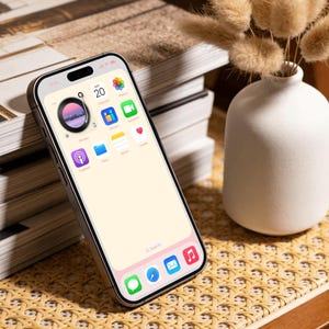 Op de afbeelding: Een zilveren smartphone met een lichtroze scherm toont app-pictogrammen. De telefoon ligt op een geweven oppervlak naast een stapel boeken en een witte vaas met gedroogde bloemen. Het scherm toont de datum als zaterdag de 20e.