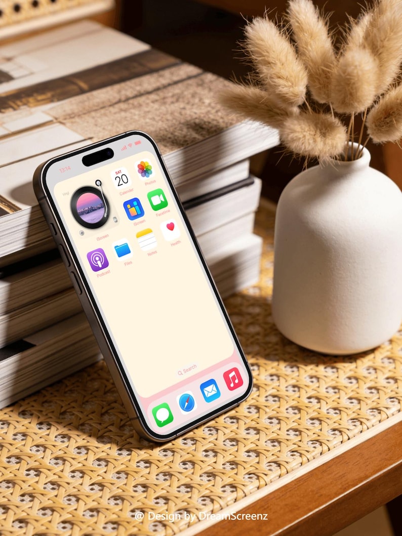 Op de afbeelding: Een zilveren smartphone met een lichtroze scherm toont app-pictogrammen. De telefoon ligt op een geweven oppervlak naast een stapel boeken en een witte vaas met gedroogde bloemen. Het scherm toont de datum als zaterdag de 20e.