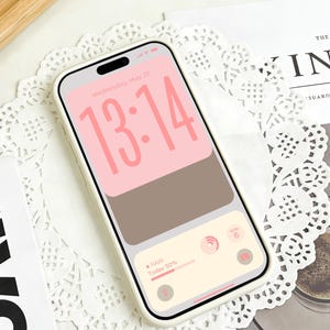 以下が含まれることがあります： 白いケースに入ったスマートフォンは、13:14の時刻と天気詳細を表示するピンクと茶色の画面を表示しています。 電話は白いレースのドイリーの上に置かれています。 テキスト「Wednesday, May 20」が表示されています。 デザインはDreamScreenzによるものです。