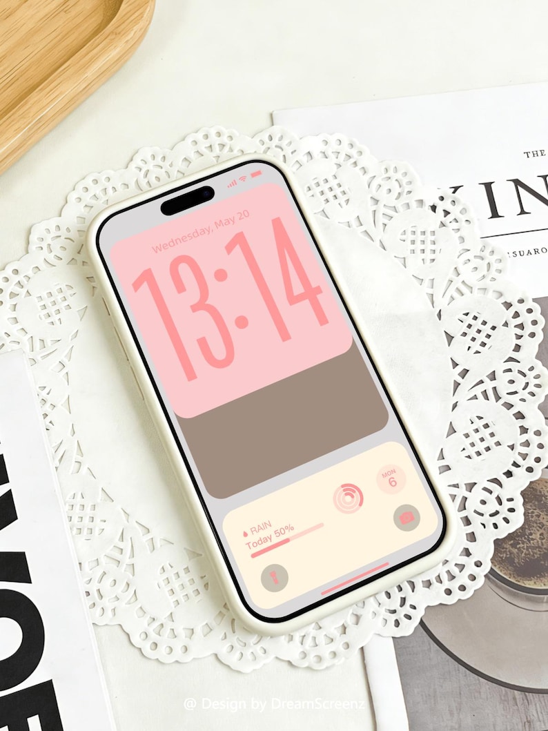 Op de afbeelding: Een smartphone met een witte hoes toont een roze en bruin scherm met de tijd 13:14. Het scherm toont ook weersinformatie. De telefoon ligt op een wit kanten kleedje. De tekst "Wednesday, May 20" is zichtbaar.