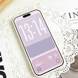 Puede incluir: Un smartphone con una funda blanca muestra una pantalla lila con la hora 13:14 y la fecha miércoles 20 de mayo. La pantalla también muestra información meteorológica. El teléfono descansa sobre un mantelito blanco de encaje.