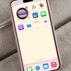 Op de afbeelding: Een roze smartphone met een zwarte rand en een lichtroze hoesje. Het scherm toont app-iconen, waaronder Agenda, Foto's en FaceTime. De achtergrond van de telefoon heeft een platenspelerontwerp. De tijd is 13:14 uur.
