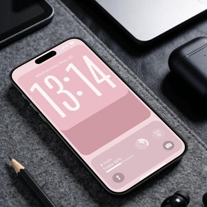 May include: A smartphone with a pink screen showing the time 13:14 and the date Wednesday, May 20. The phone is on a grey surface with a pencil, laptop, and wireless earbuds. The screen displays a weather forecast of 50% chance of rain.