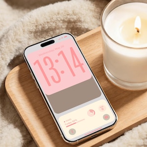Op de afbeelding: Een smartphone met een gouden frame toont een roze en bruin scherm met de tijd 13:14. Het ligt op een houten dienblad naast een brandende kaars in een glas. De achtergrond is een zachte, witte, getextureerde stof. De tekst "@ Design by DreamScreenz" staat onderaan.