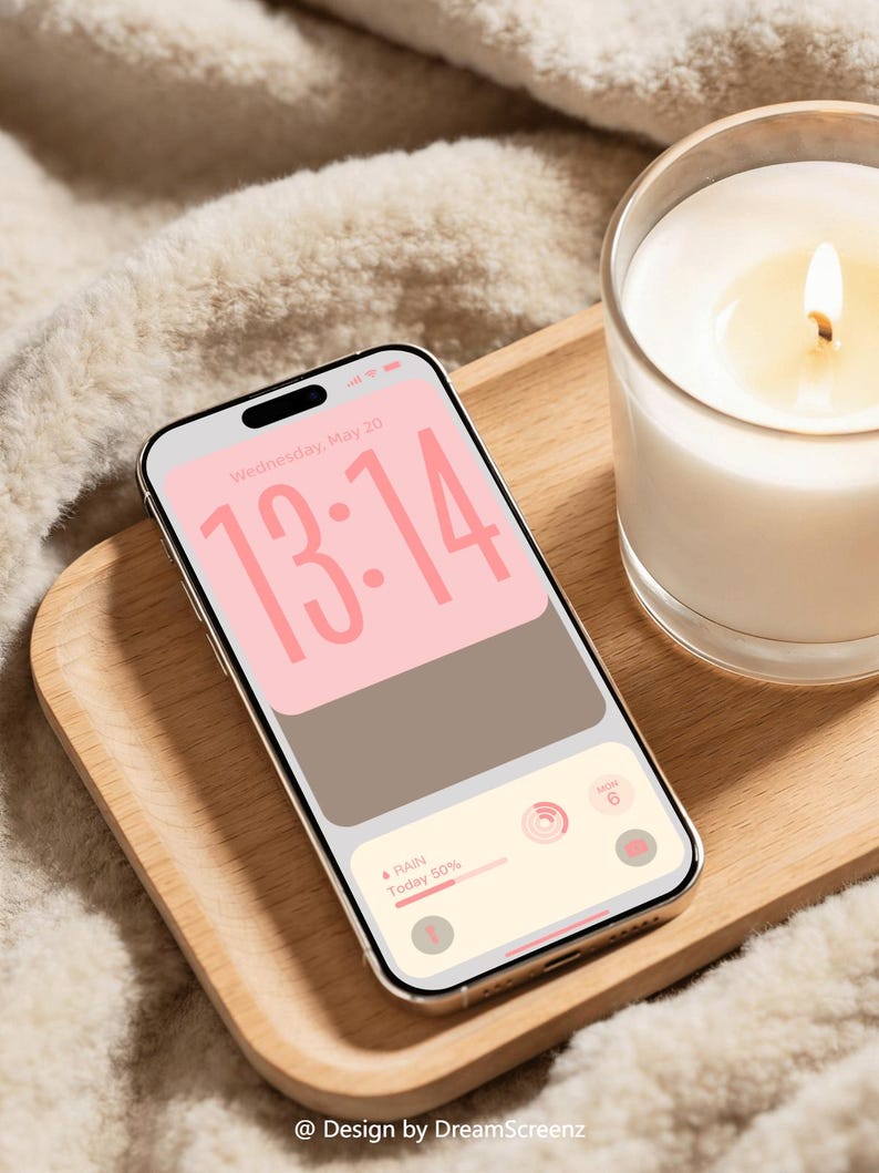 Op de afbeelding: Een smartphone met een gouden frame toont een roze en bruin scherm met de tijd 13:14. Het ligt op een houten dienblad naast een brandende kaars in een glas. De achtergrond is een zachte, witte, getextureerde stof. De tekst "@ Design by DreamScreenz" staat onderaan.