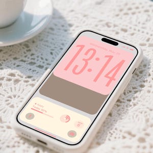 Op de afbeelding: Een witte smartphone in een witte hoes toont een digitale klok met 13:14 in roze. Het scherm toont ook een weerwidget met 50% kans op regen. Een kopje koffie staat op een witte schotel op de achtergrond.