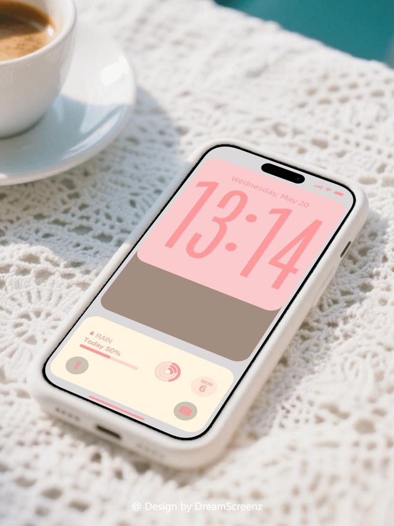 Op de afbeelding: Een witte smartphone in een witte hoes toont een digitale klok met 13:14 in roze. Het scherm toont ook een weerwidget met 50% kans op regen. Een kopje koffie staat op een witte schotel op de achtergrond.