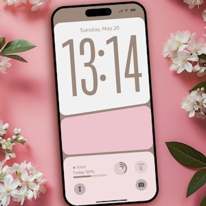 Puede incluir: Un teléfono inteligente con una pantalla rosa claro y beige que muestra la hora 13:14, la fecha y la información meteorológica. Flores blancas y hojas verdes están dispuestas alrededor del teléfono sobre un fondo rosa. El texto "@ Created by DreamScreenz" está en la parte inferior.