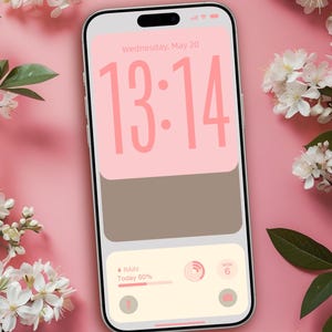 Op de afbeelding: Een smartphonescherm toont de tijd 13:14 in roze, met een roze en bruin kleurenschema. Het scherm toont ook de datum, weersinformatie en een camera-icoon. Witte bloemen en groene bladeren zijn rond de telefoon gerangschikt op een roze achtergrond. De tekst "@ Design by DreamScreenz" staat onderaan.