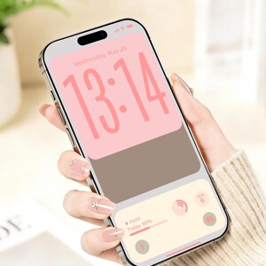 Op de afbeelding: Een zilveren smartphone toont een roze en bruine digitale klok met de tijd 13:14. Het scherm toont ook de datum, het weer en een kalenderpictogram. De telefoon wordt vastgehouden in een hand met verzorgde nagels.
