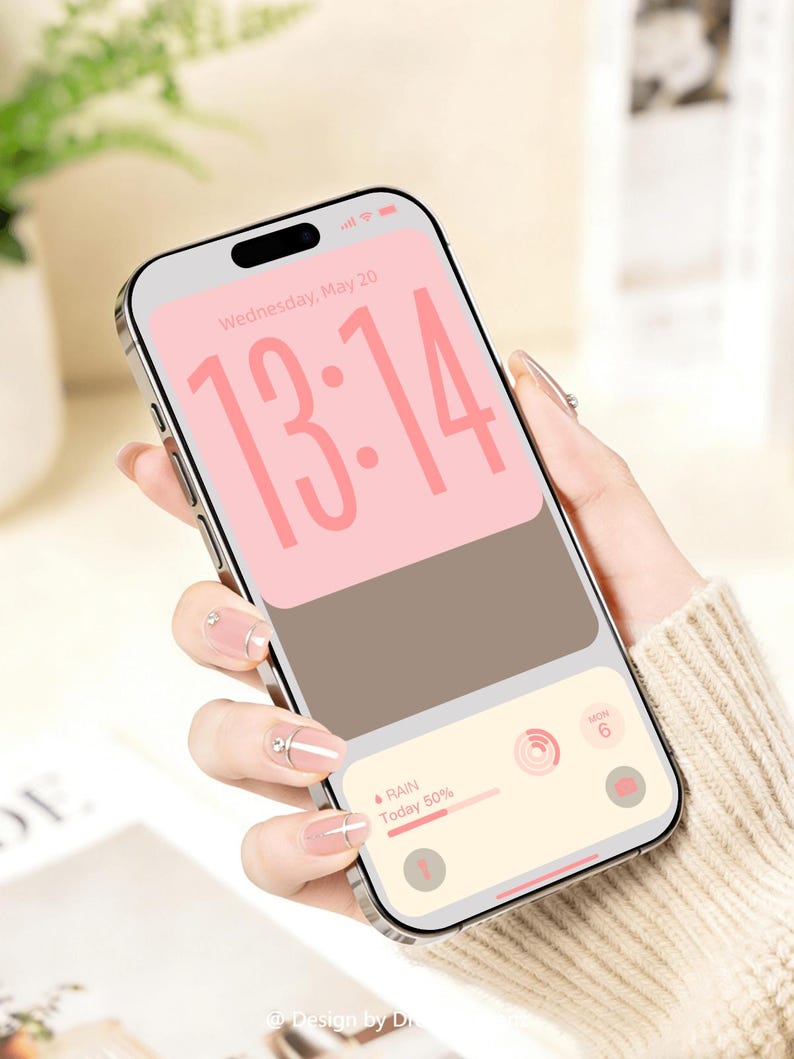 Op de afbeelding: Een zilveren smartphone toont een roze en bruine digitale klok met de tijd 13:14. Het scherm toont ook de datum, het weer en een kalenderpictogram. De telefoon wordt vastgehouden in een hand met verzorgde nagels.