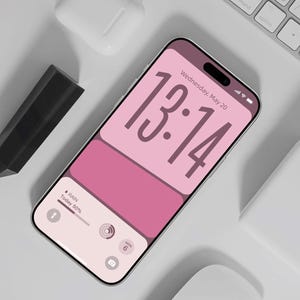 以下が含まれることがあります： 13時14分と5月20日水曜日を表示するピンク色の画面のスマートフォン。スマートフォンの周りには、白いキーボード、白いマウス、白い充電ケース、黒いスタイラスがあります。画面には雨50％も表示されています。