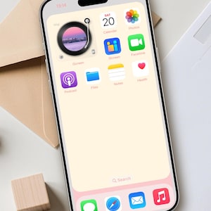 Op de afbeelding: Een smartphone scherm toont een aangepast startscherm met een vinyl record widget, kalender en app iconen. De achtergrond is een zachte perzikkleur met een roze rand. De tijd is 13:14. Design by DreamScreenz.