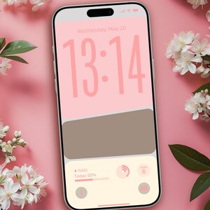 Puede incluir: Un teléfono inteligente muestra una pantalla de bloqueo personalizada con un fondo rosa y grandes números rosas que indican la hora, 13:14. La pantalla también muestra un pronóstico del tiempo y un widget rectangular marrón. Flores blancas y hojas verdes están dispuestas alrededor del teléfono.