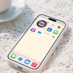Op de afbeelding: Een witte smartphone met een scherm met app-pictogrammen, waaronder een kalender met de datum 20. De telefoon zit in een witte hoes en ligt op een wit, gestructureerd oppervlak. Een kopje koffie staat op de achtergrond.