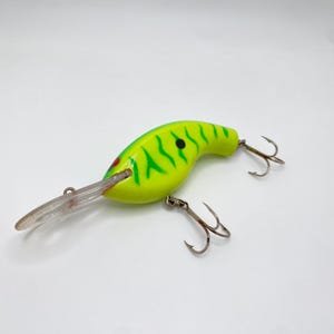 May include: A bright yellow fishing lure with green stripes and a red eye. The lure has two treble hooks and a clear plastic lip. This fishing lure is designed to attract fish.