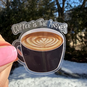 Peut inclure: Un autocollant transparent représentant une tasse de café noire avec un motif de latte et le texte "Coffee is the Answer" en blanc. Le café a un motif en forme de cœur dans la mousse. L'autocollant est tenu devant un fond enneigé.