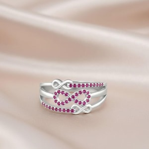 Könnte beinhalten: Ein silberner Ring mit drei Bändern, jedes mit einem Unendlichkeitssymbol. Die Unendlichkeitssymbole sind mit kleinen, runden, rosa Edelsteinen verziert. Der Ring ist vor einem weichen, rosa Hintergrund platziert.